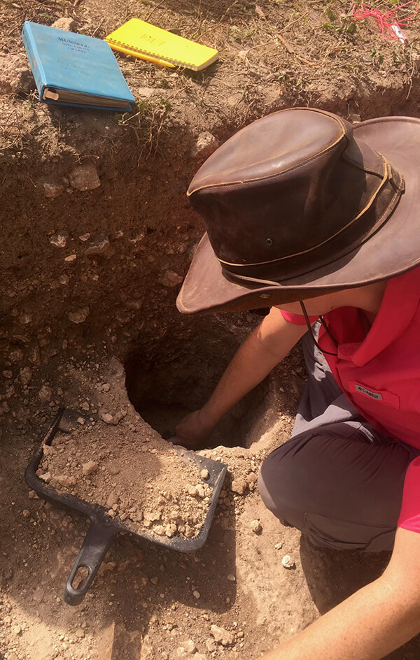 A person wearing a hat digs into a hole in the ground with a binder, pen and notepad in the background